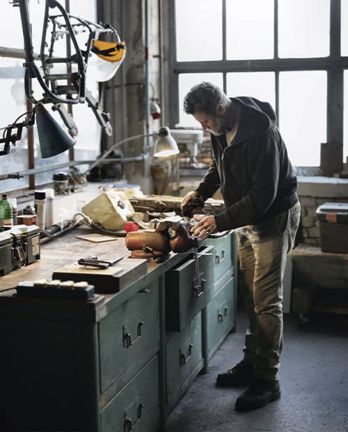 Jouw dagbesteding, jouw nieuwe plek. Een man werkt aan een rommelige werkbank in een goed verlichte werkplaats, omringd door gereedschap en apparatuur.