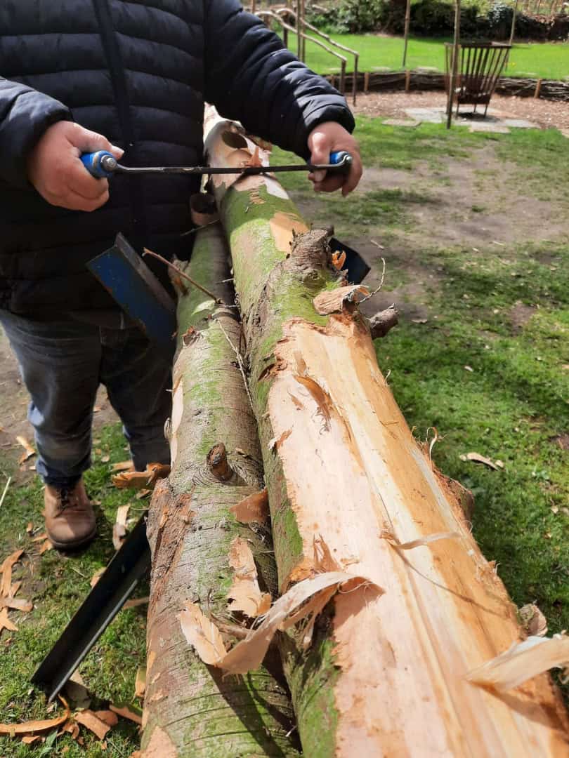 Jouw dagbesteding, jouw nieuwe plek. Een persoon die buiten met gereedschap de bast van een gevallen boomstam afpelt.